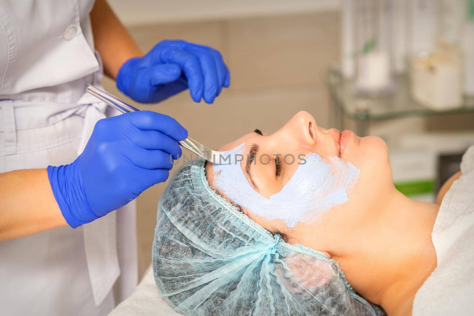 The beautician with brush applies a photochemical and glycolic peeling face mask to the female patient face in the beauty clinic. by okskukuruza