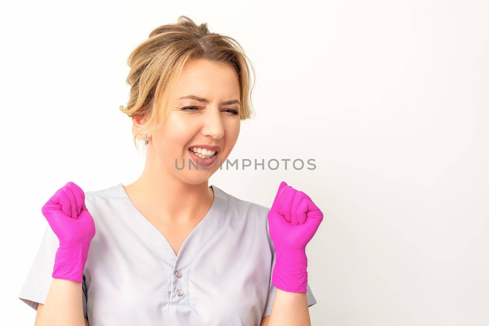 Happy caucasian woman doctor wearing pink gloves celebrates and raising fists on white background