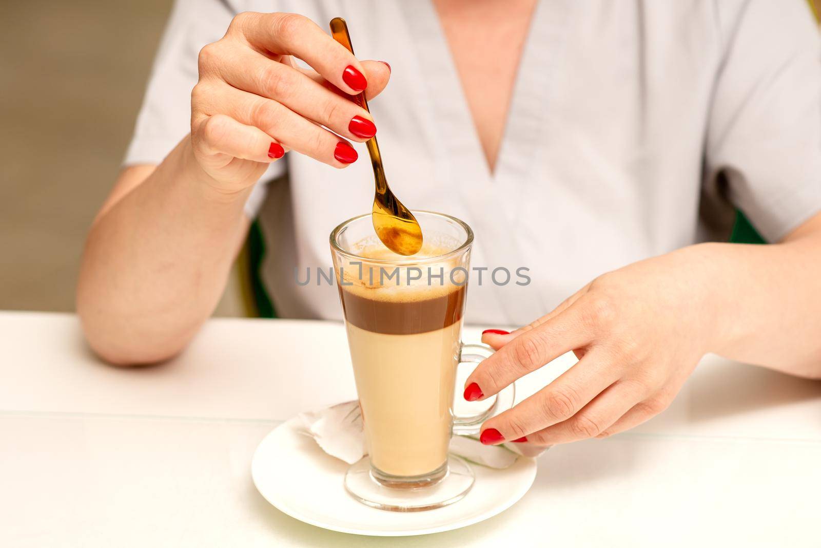 Woman with the latte. Glass mug of latte coffee on the white saucer with female hands holding teaspoon on the table in the cafeteria, close up. by okskukuruza