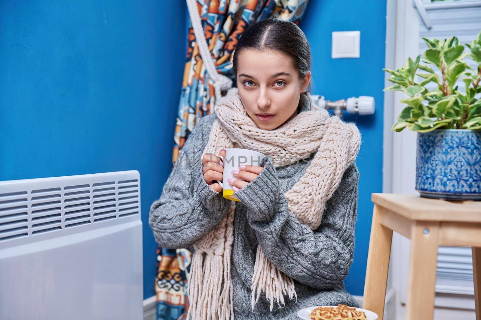 Cold autumn winter season, lifestyle, teenage female in warm sweater scarf warming near electric heating radiator, drinking hot drink with cup. Heating, warmth, equipment, life in cold season