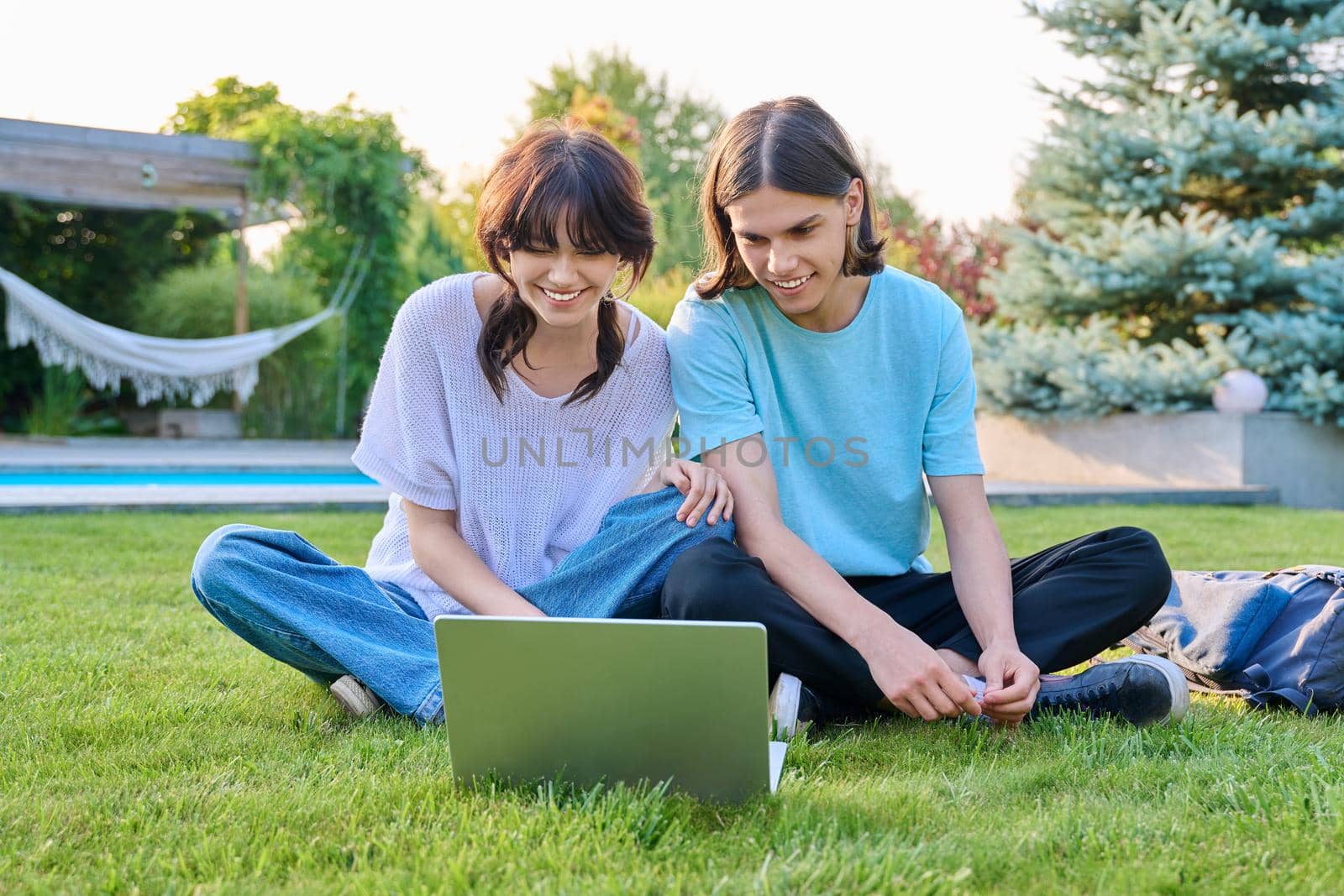 Two teenage friends of students sitting on grass with laptop, in backyard, guy and girl 18 years old study together. Friendship, youth, technology, high school, college, lifestyle concept