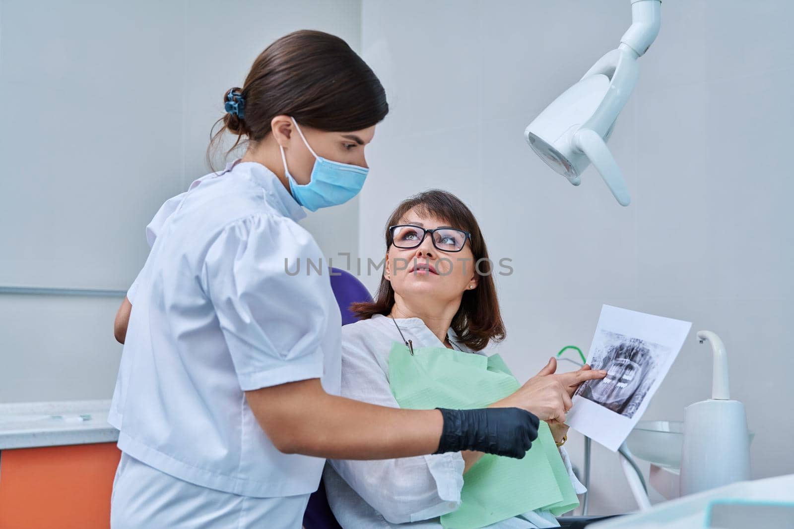 Female doctor dentist talking to middle aged woman patient in dental chair, discussing x-rays of teeth and jaws. Dental treatment and prosthetics, implantation, health care concept