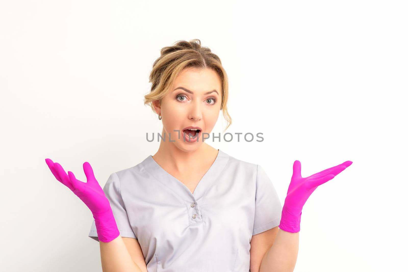 Body language. Young surprised Caucasian woman doctor wearing gloves gesturing with her hands, spread his arms having shocked expression opening her mouth against the white background