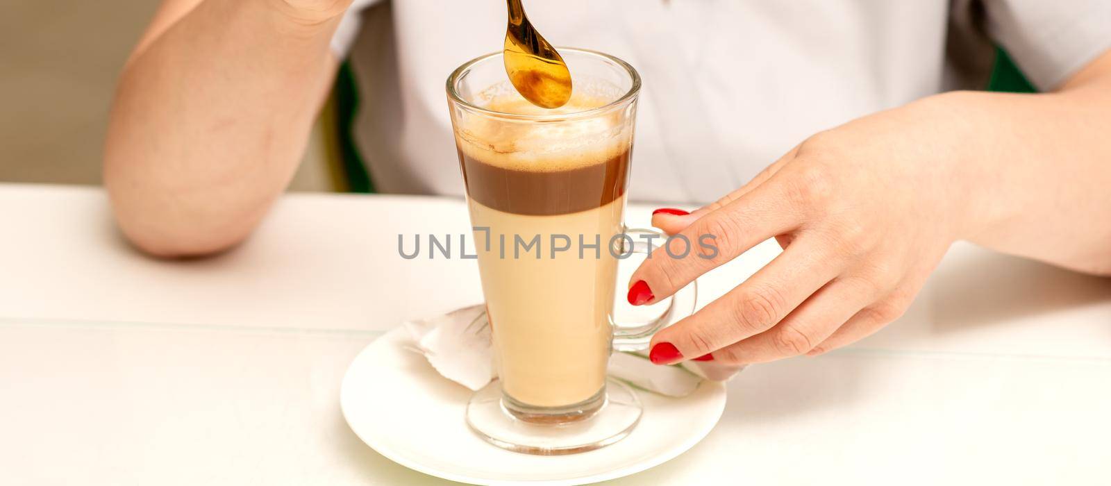 Woman with the latte. Glass mug of latte coffee on the white saucer with female hands holding teaspoon on the table in the cafeteria, close up. by okskukuruza