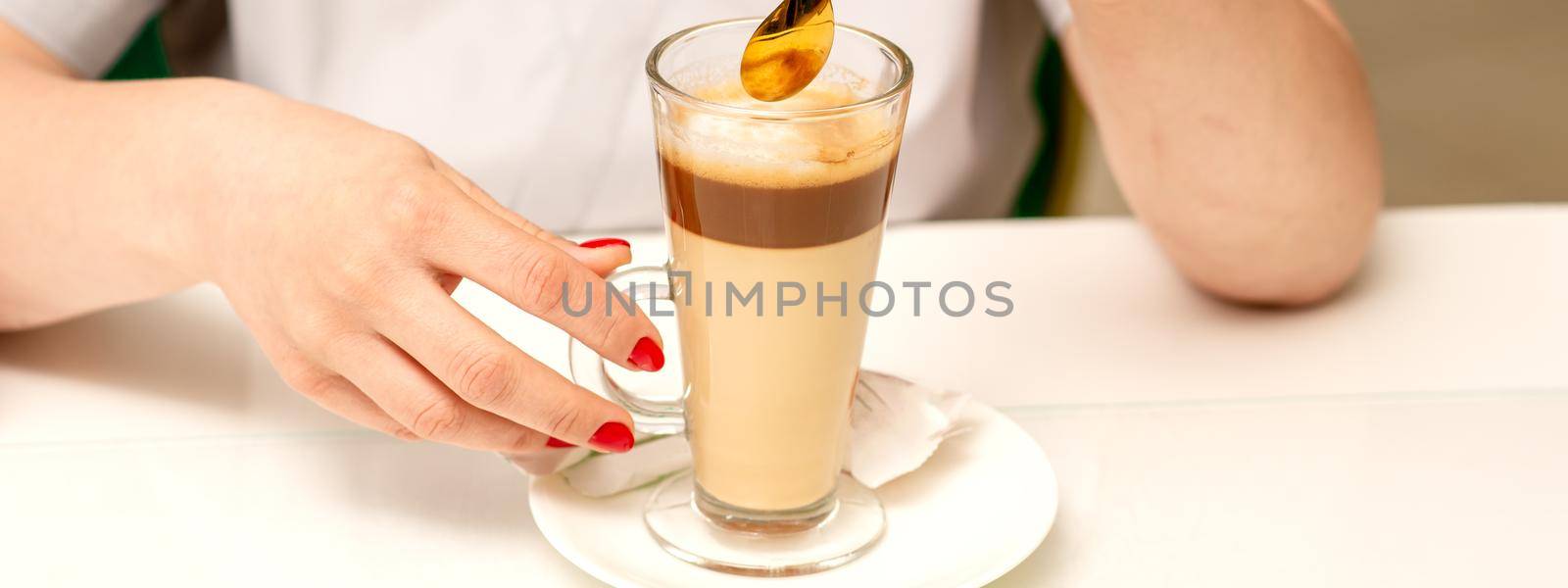 Woman with the latte. Glass mug of latte coffee on the white saucer with female hands holding teaspoon on the table in the cafeteria, close up