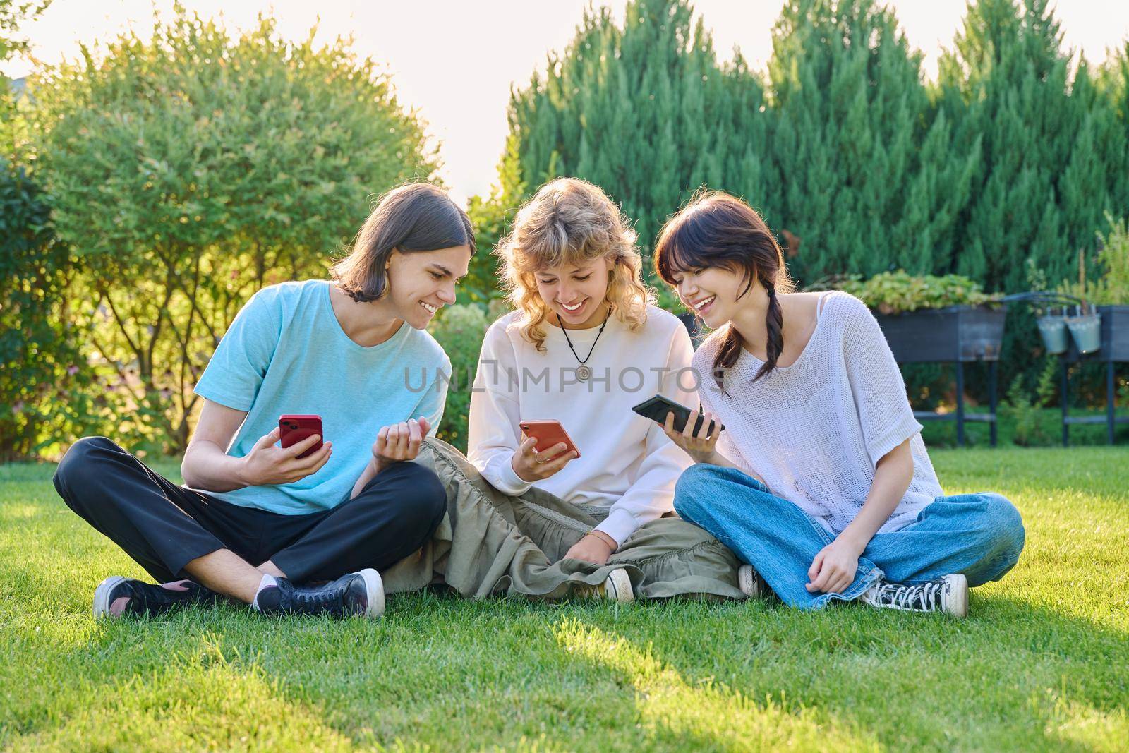 Group of teenagers sitting on grass with smartphones. Three teenage friends relaxing on lawn, sunny summer day, using mobile applications for leisure. Lifestyle technology and friendship youth concept