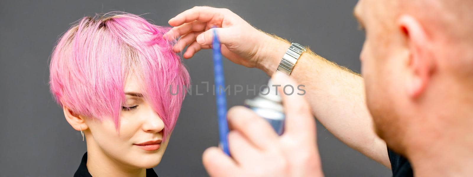 The hairdresser is using hair spray to fix the short pink hairstyle of the young caucasian woman in the hair salon