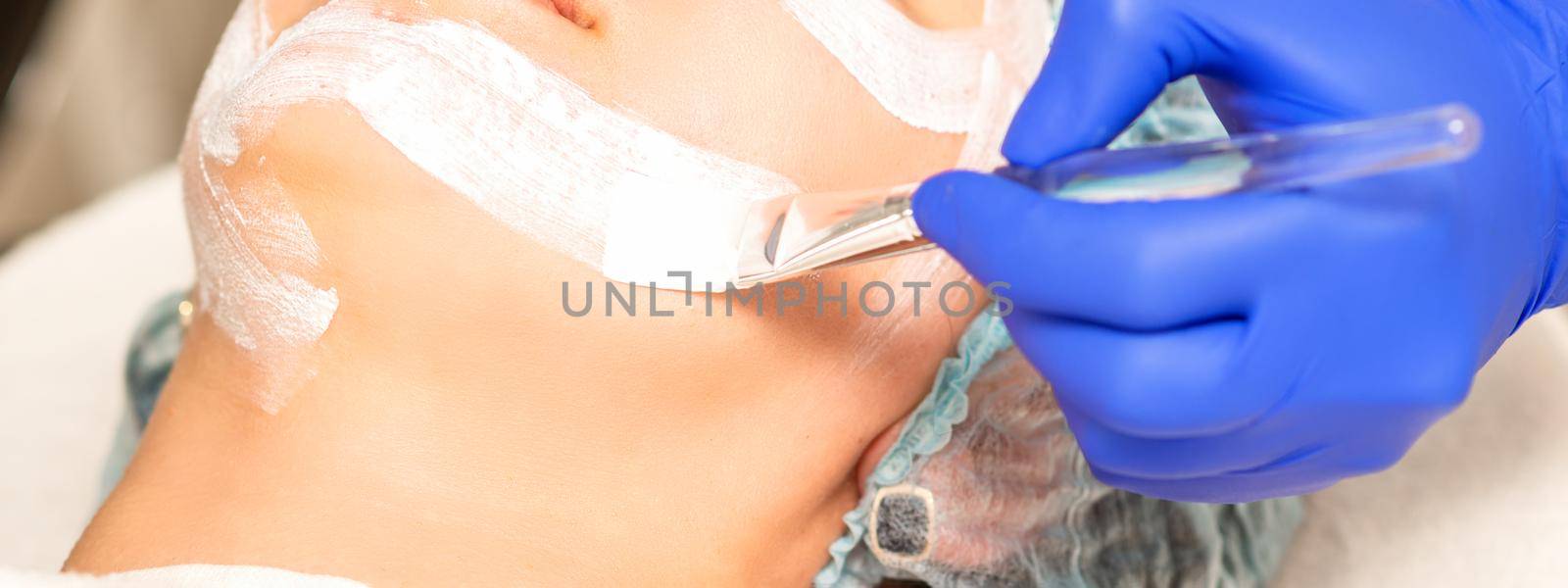 The beautician with brush applies a photochemical and glycolic peeling face mask to the female patient face in the beauty clinic