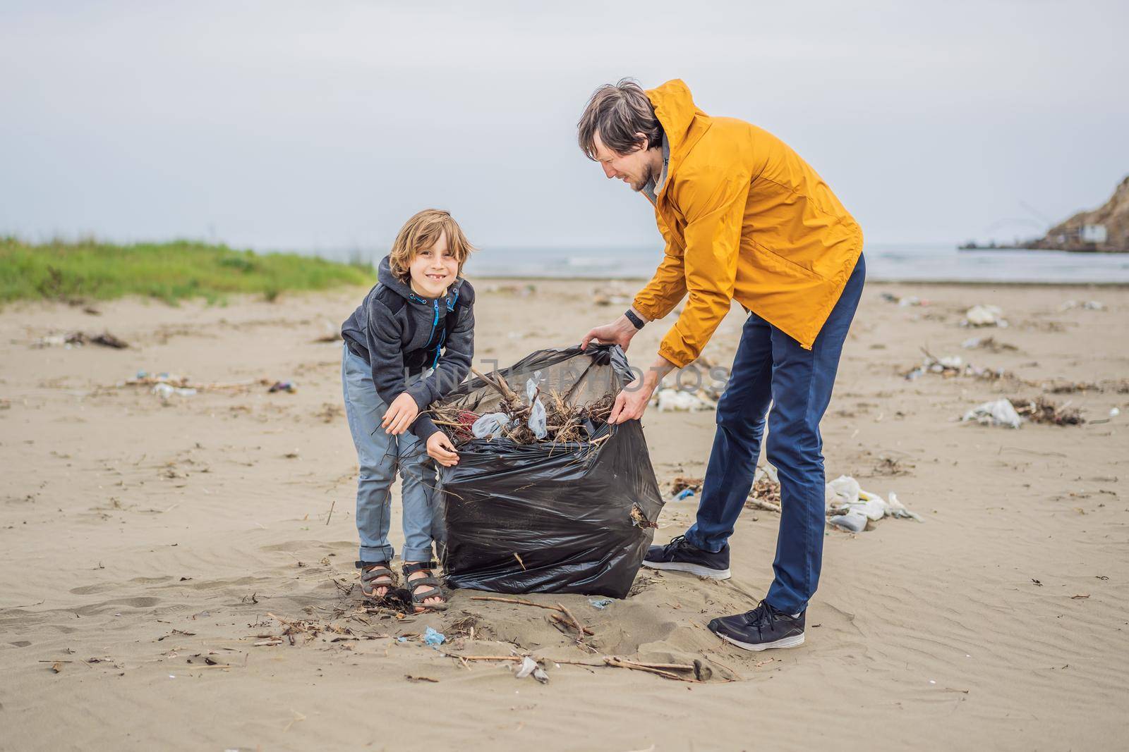 Dad and son in gloves cleaning up the beach pick up plastic bags that pollute sea. Natural education of children. Problem of spilled rubbish trash garbage on the beach sand caused by man-made by galitskaya