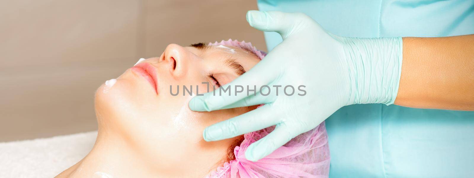 Cosmetologist with gloved hands applies a moisturizing mask with peeling cream on the female face. Facial cosmetology treatment. Procedures for facial care