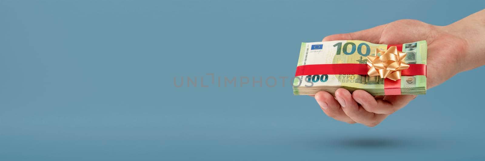 Gift of money. A man holds a stack of banknotes in his hands as a symbol or concept of a gift. 100 euro banknotes tied with a red ribbon with a bow on a blue background