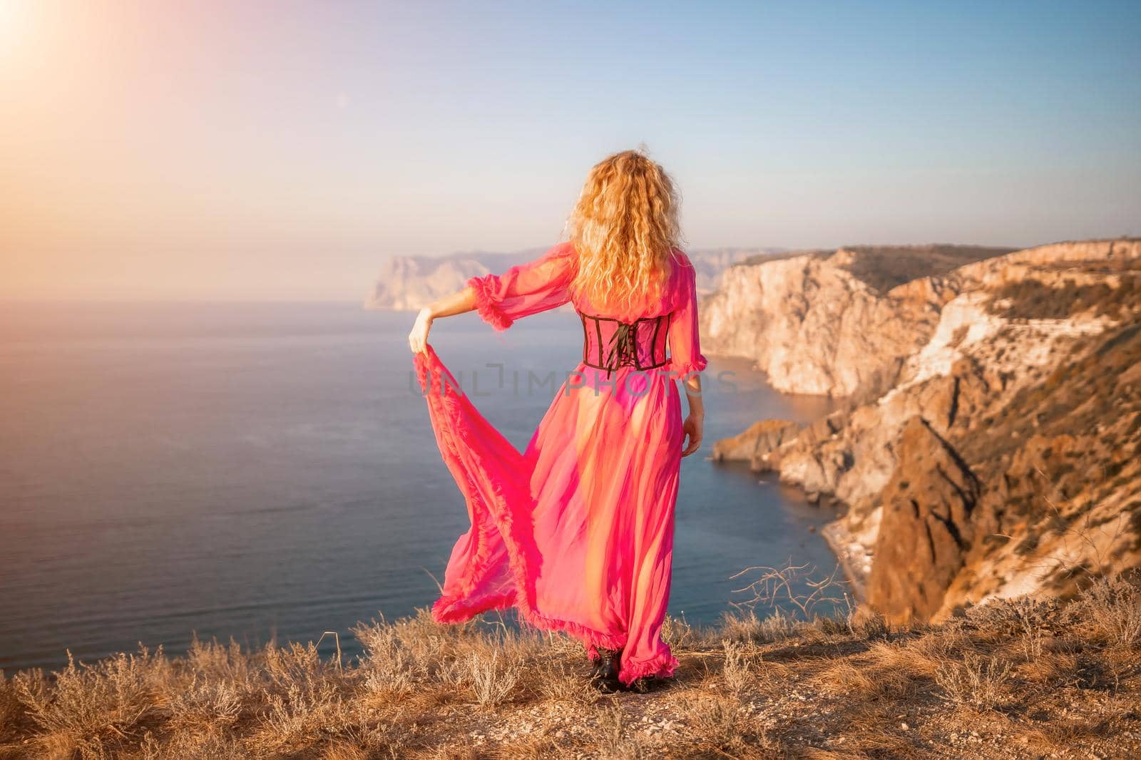 Side view a Young beautiful sensual woman in a red long dress posing on a volcanic rock high above the sea during sunset. Girl on the nature on blue sky background. Fashion photo