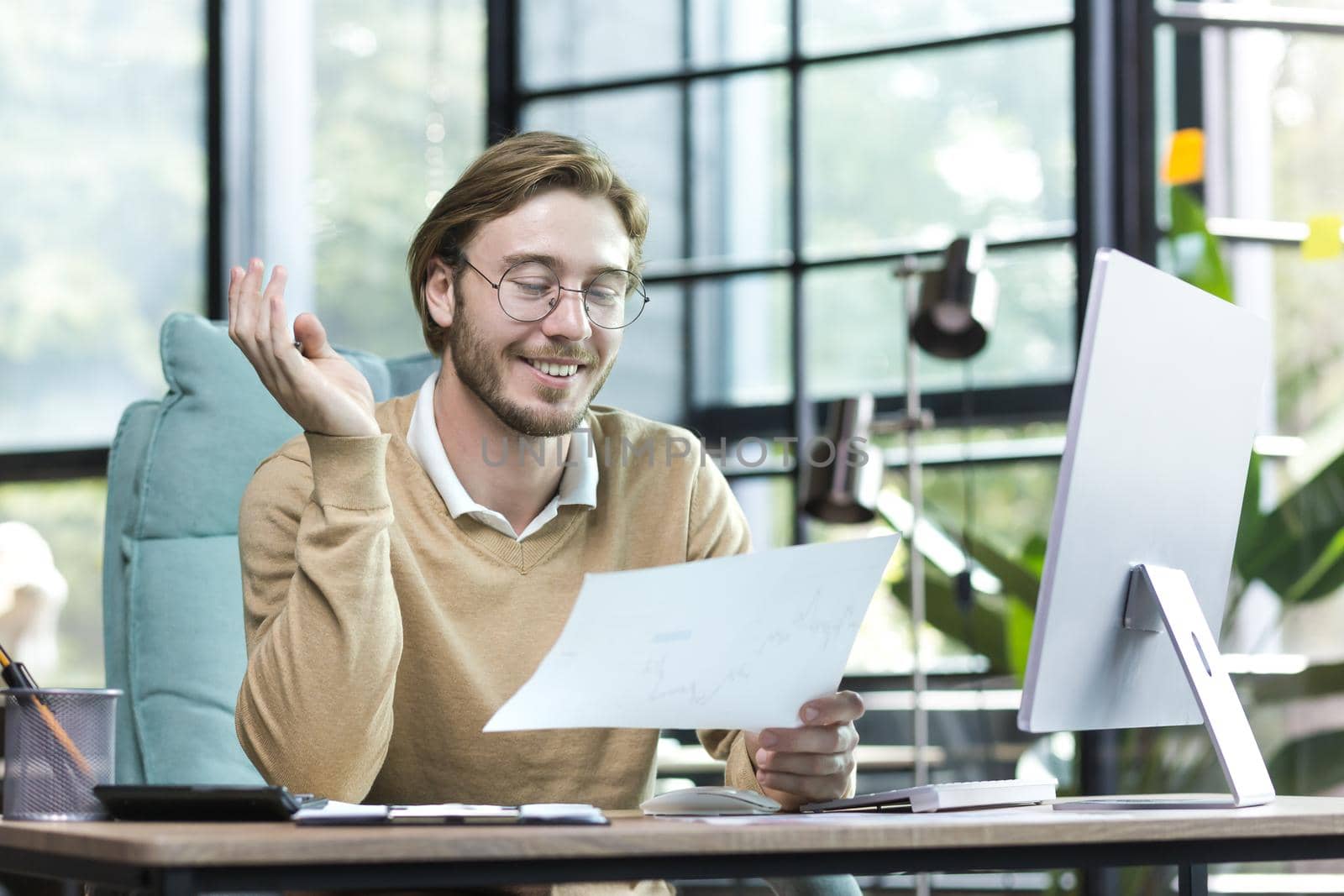 Happy blond man working inside an office building, young financier working with documents on paper work, businessman at computer smiling and reading financial report
