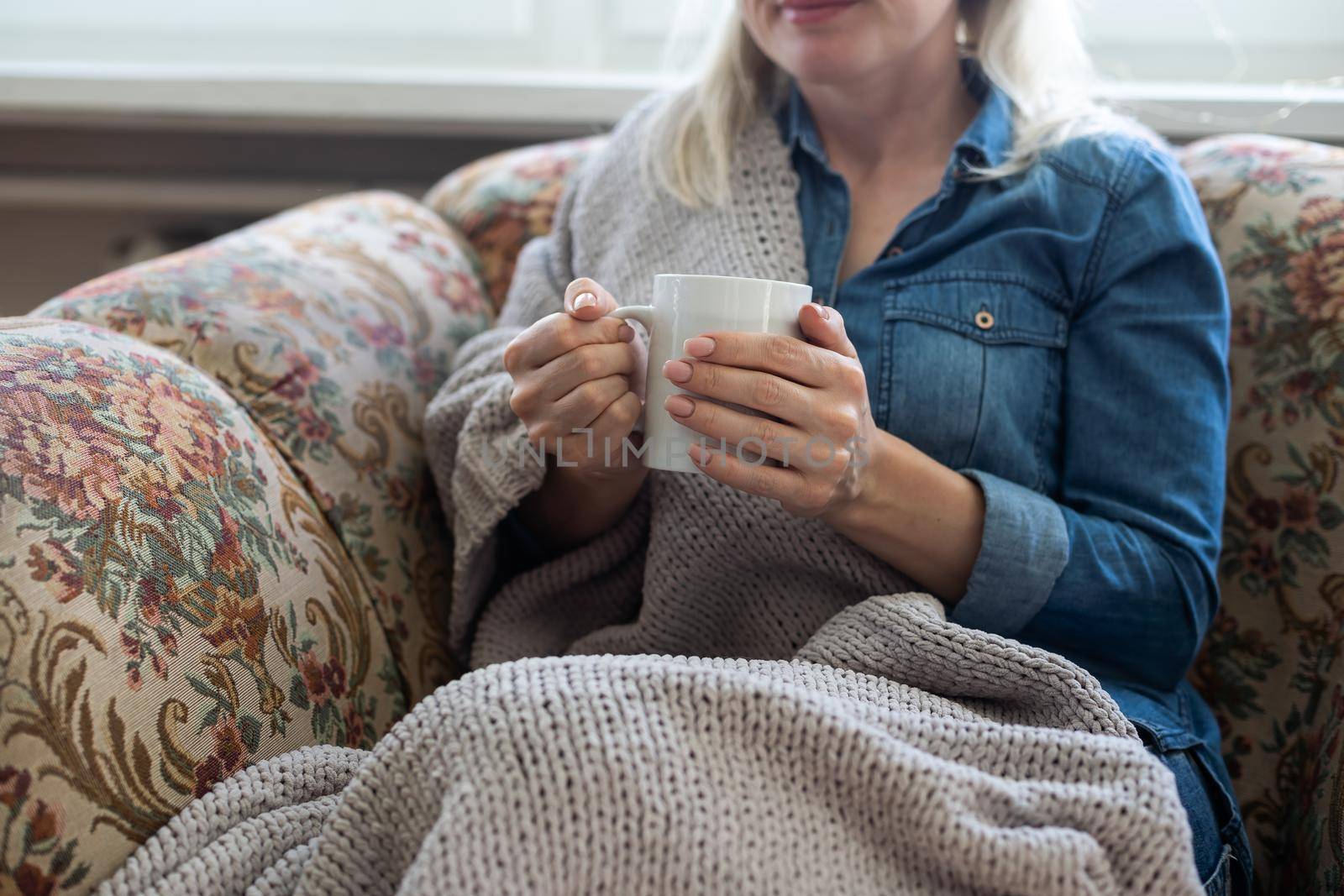 Close up of hands holding blue cup of tea or coffee. Model and cozy plaid is sitting, relax at home on sofa.