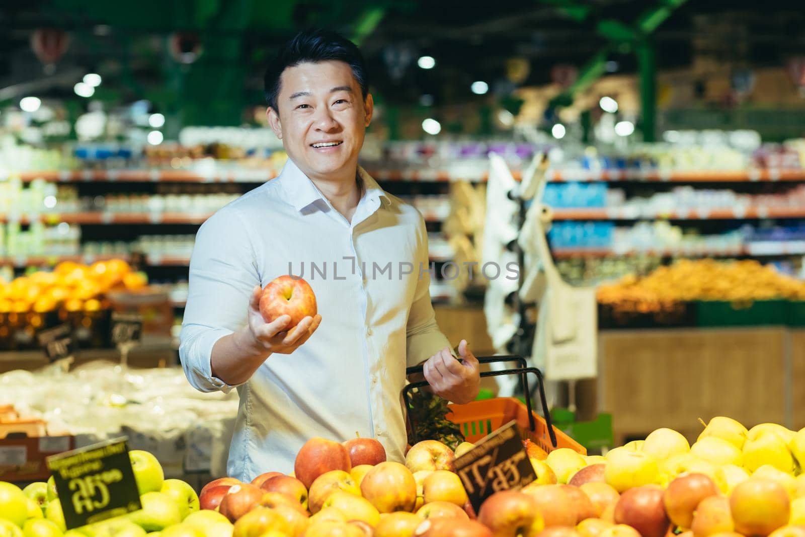 A handsome young Asian man is shopping in a supermarket, standing with a grocery basket near the fruit and vegetable section, holding an apple in his hand, looking at the camera and smiling.