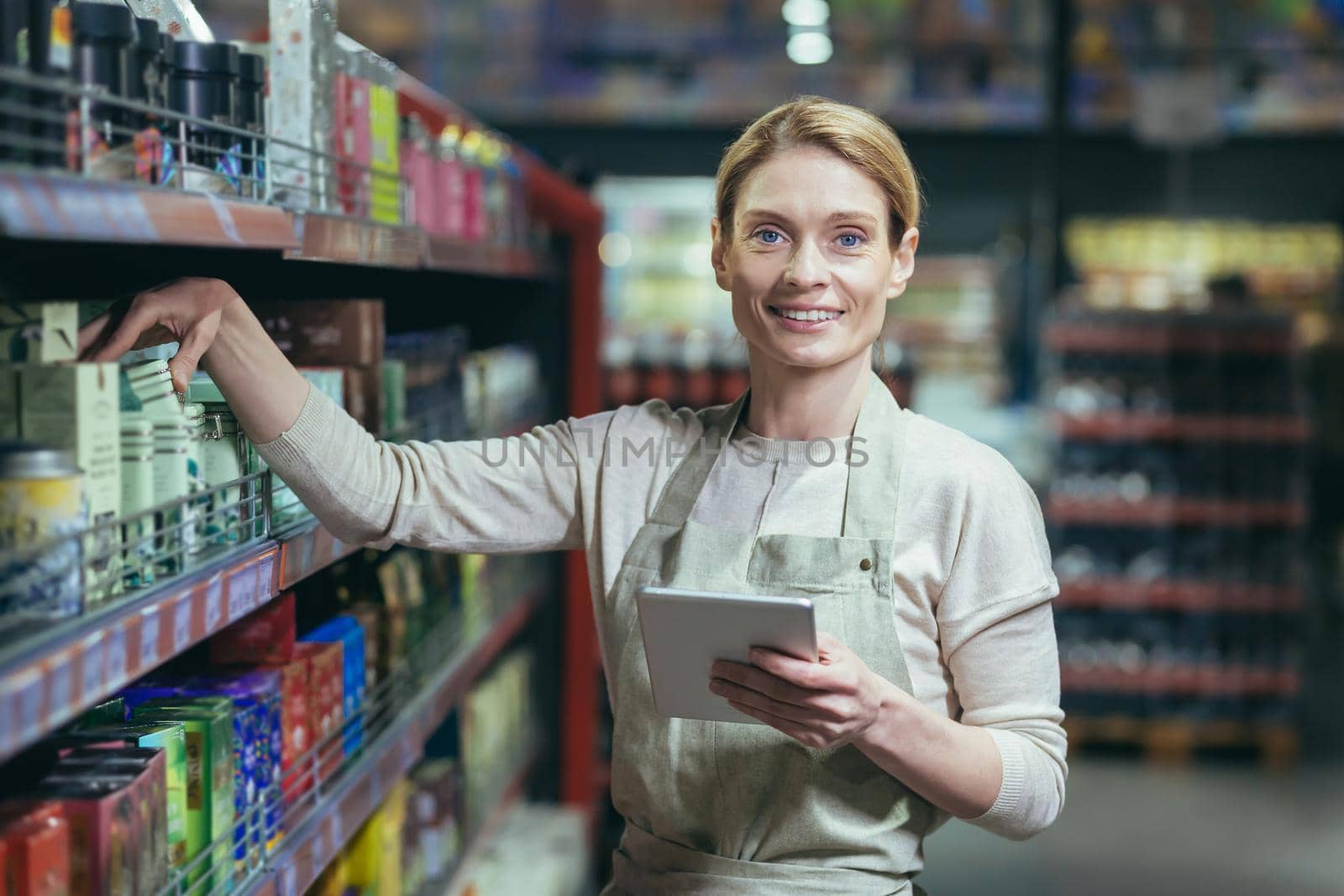 A young beautiful business woman, the owner of a store, a supermarket, stands in an apron near the shelves with products, holds a notebook in her hands, looks at the camera, smiles.