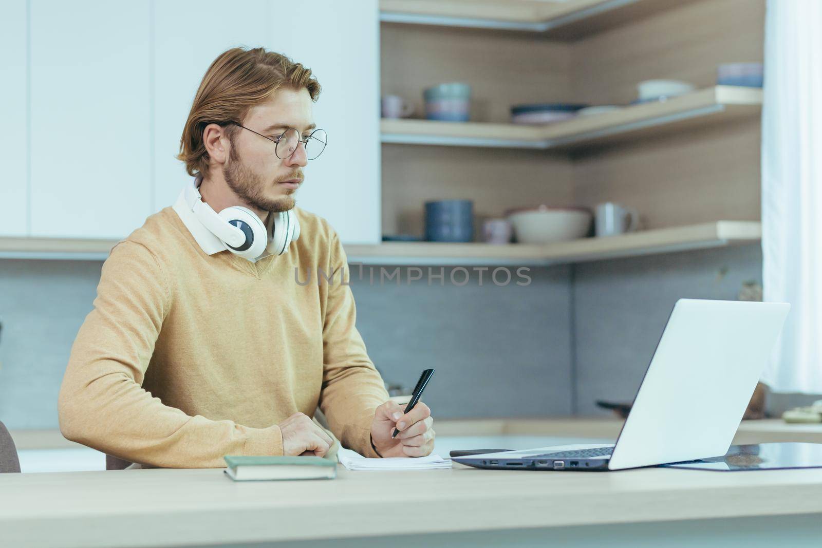 Young serious and focused man studying remotely at home, student in kitchen wearing glasses using laptop for distance learning taking notes on study material.