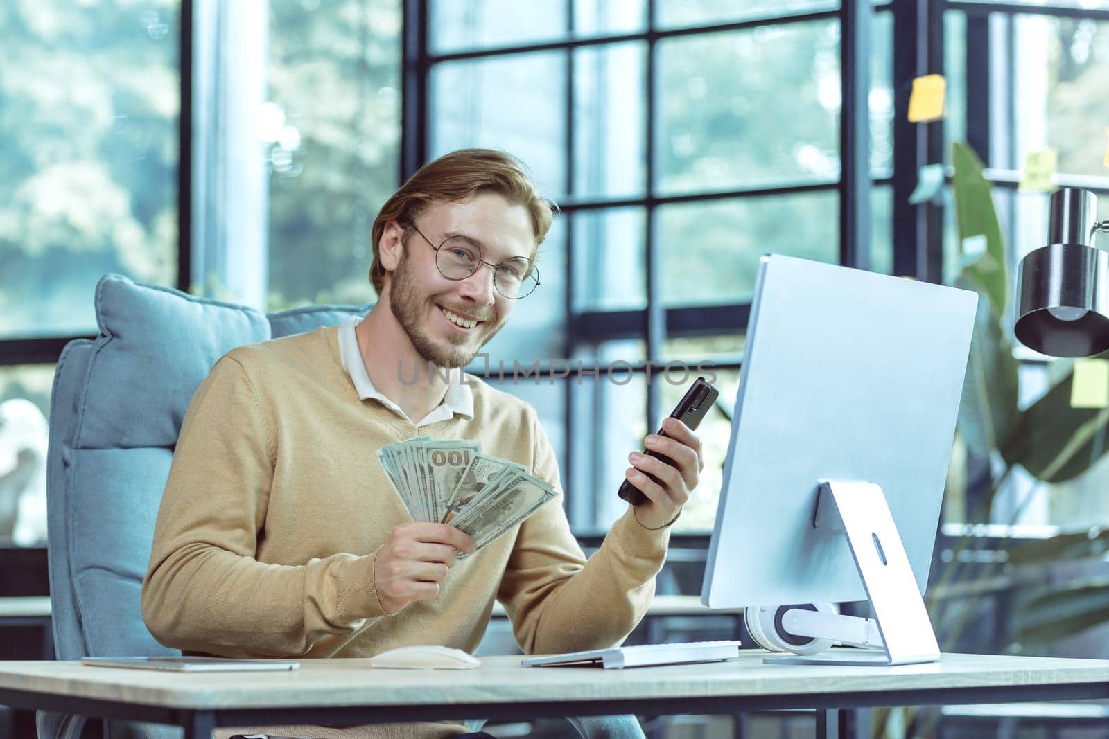 Portrait of successful financial investor, businessman smiling and looking at camera man holding dollars cash and mobile phone working inside modern office.
