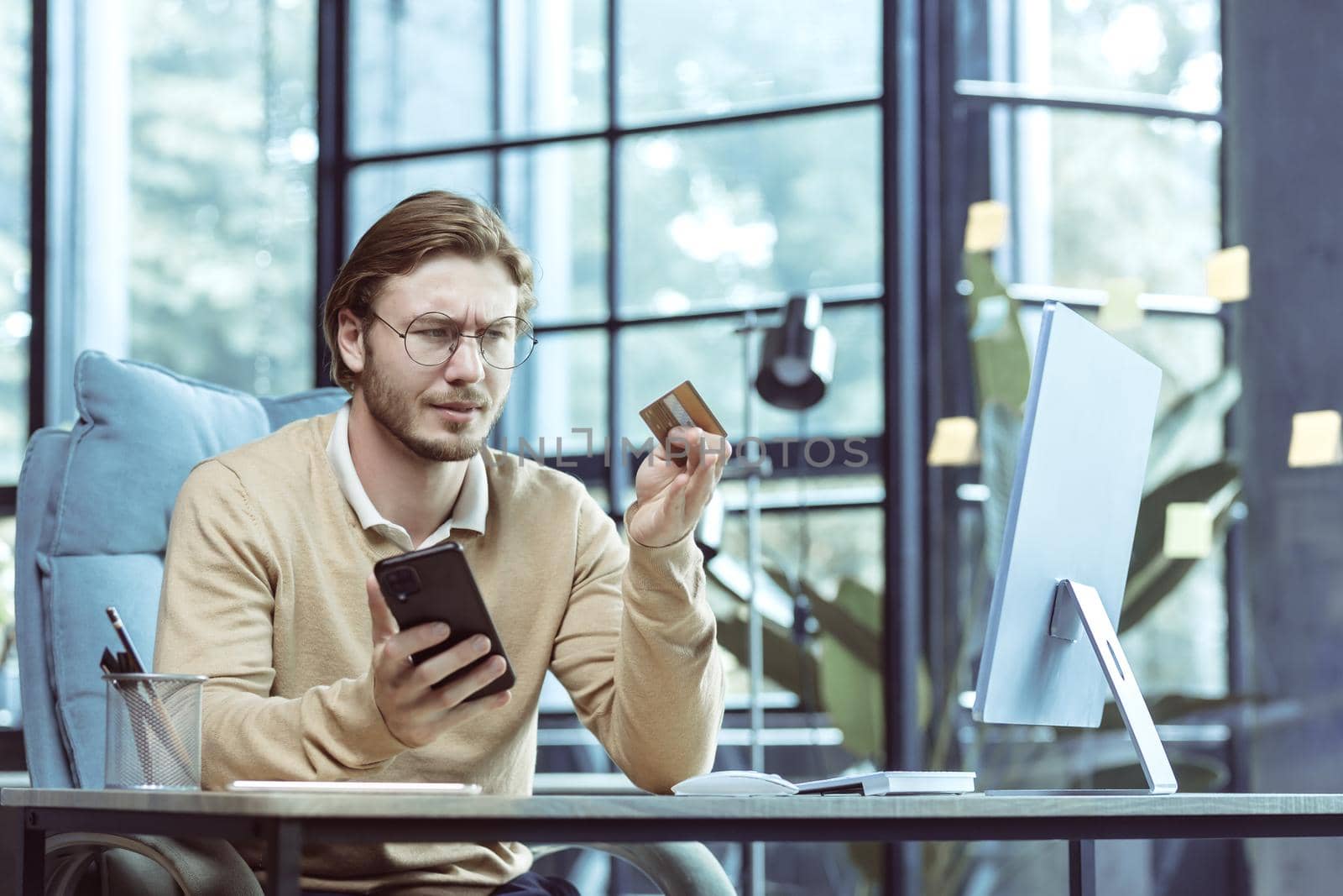 Frustrated and unsatisfied businessman working in modern loft office, freelancer cheated and trying to make bank transaction, using bank credit card and smartphone