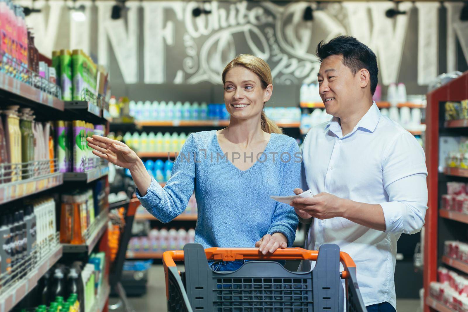 A young international couple, a woman and an Asian man walk between rows with a shopping cart in a supermarket, the woman points to the product, the man holds a shopping list, smilling.