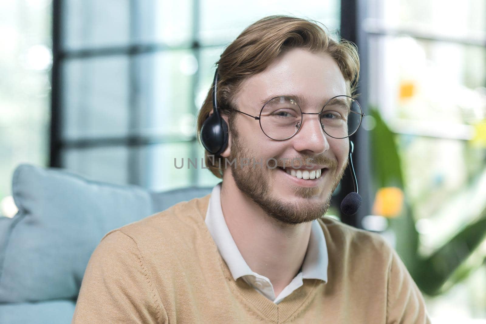 Close-up photo of man wearing headset for video call, businessman working inside office building, smiling and looking at camera, employee at work wearing glasses