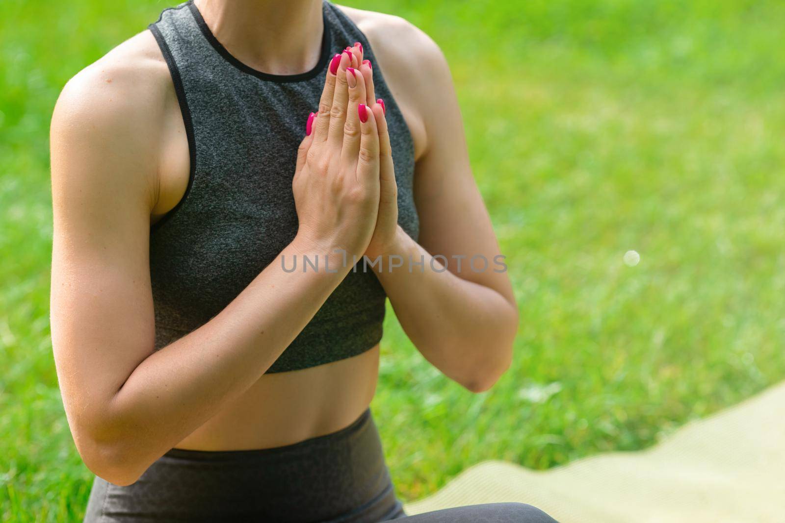 A close-up of some slender woman in a gray top, sitting on the green grass in the park, with her palms folded in front of her. Copy space
