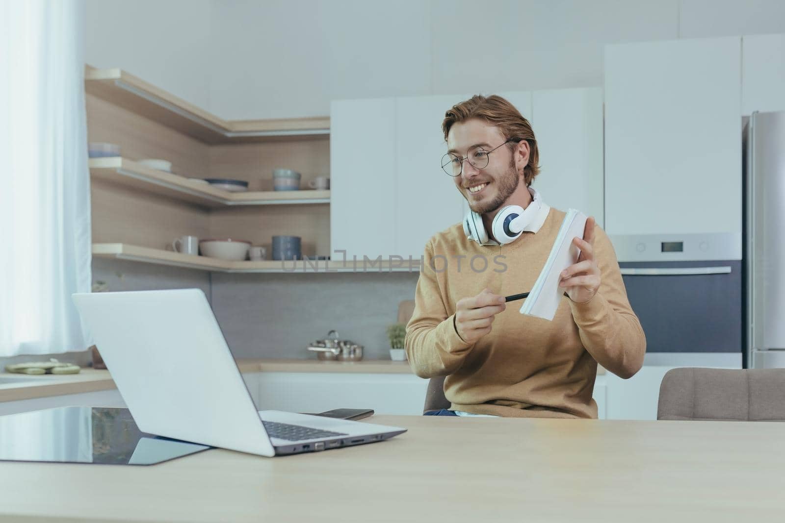 Young teacher teaches remotely from home, man works in kitchen at home, uses laptop for online call with communicates with students smiling explains assignment shows notebook.