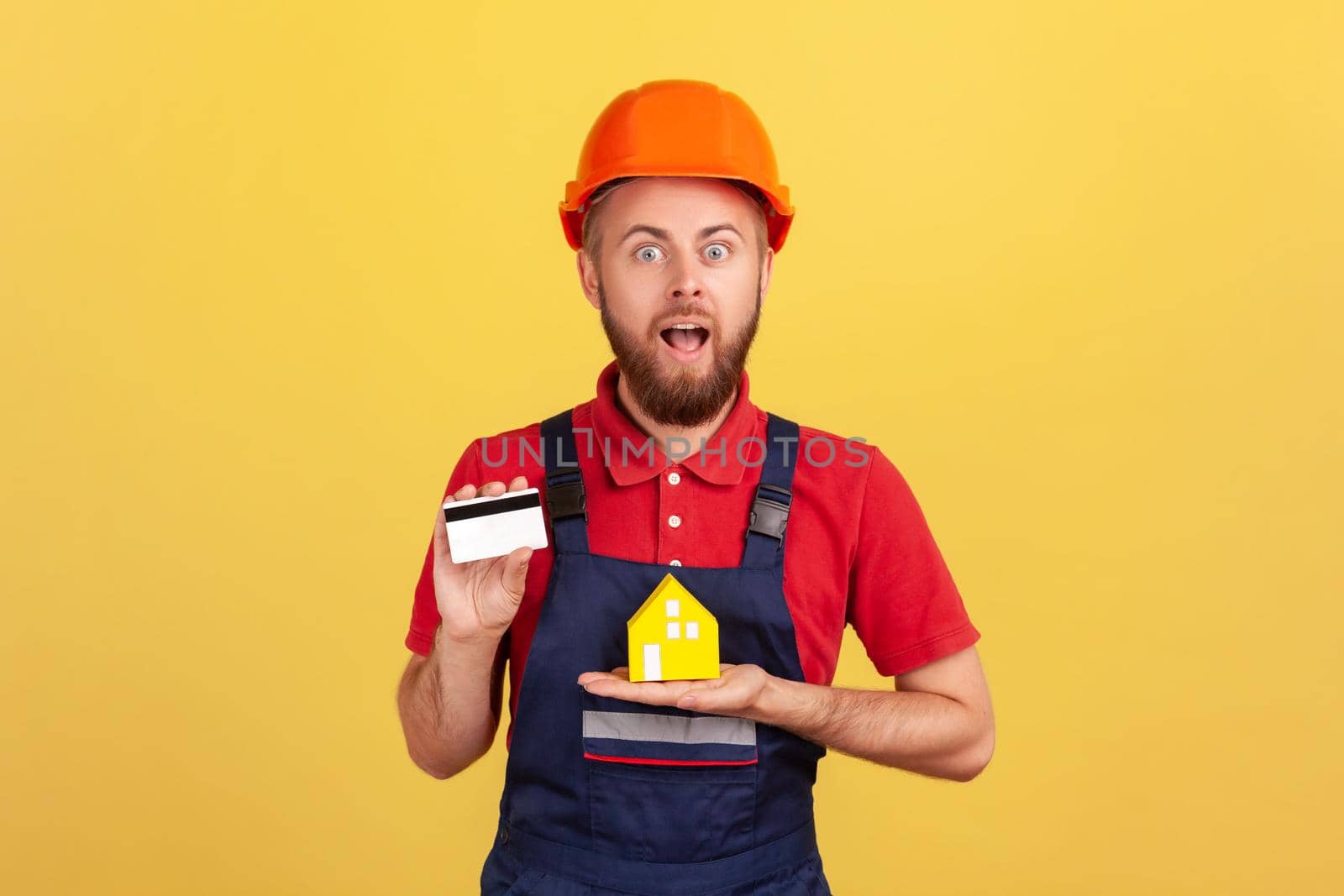 Portrait of shocked builder man wearing blue uniform and protective helmet showing credit card and paper house, looking at camera with open mouth. Indoor studio shot isolated on yellow background.