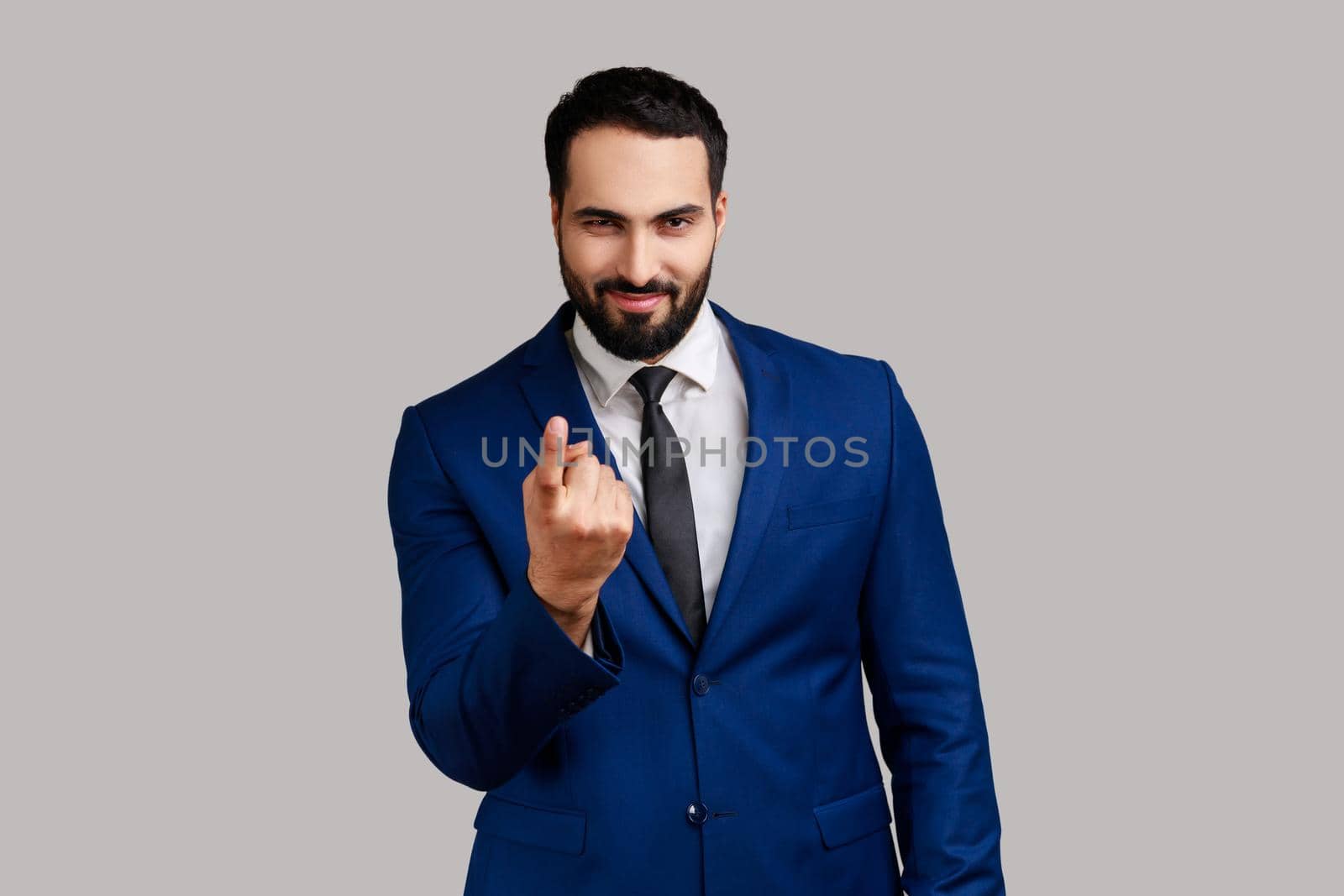 Portrait of positive smiling handsome bearded businessman calling with finger gesture, inviting to come in, wearing official style suit. Indoor studio shot isolated on gray background.