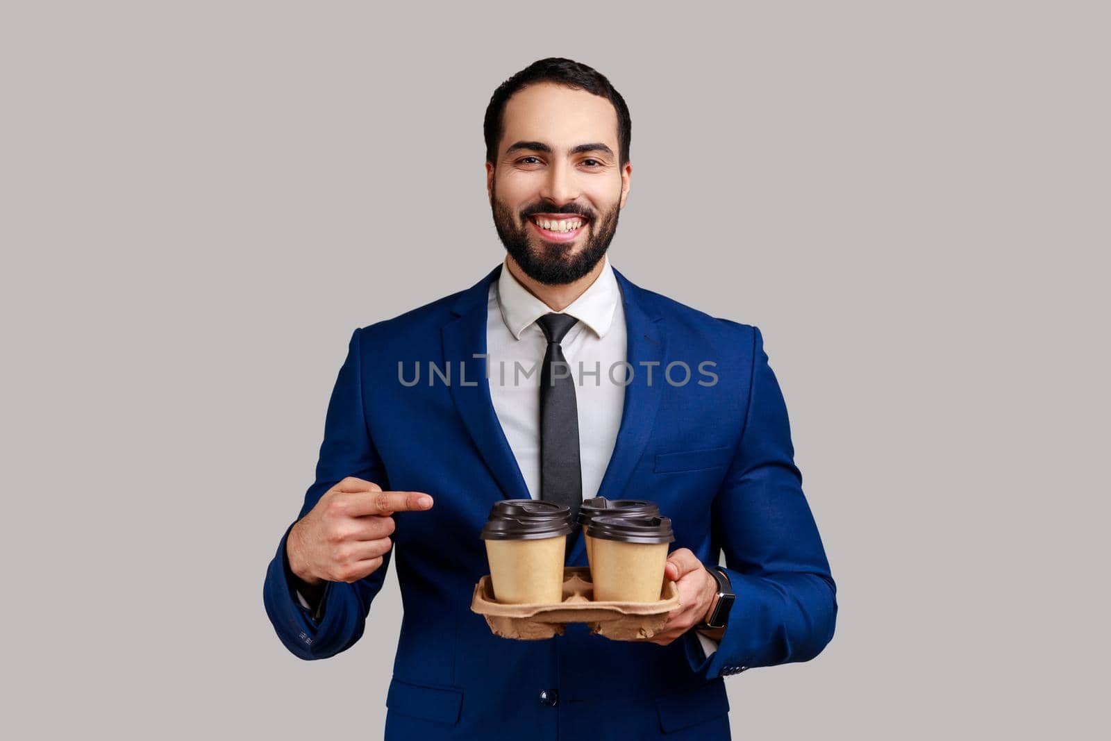 Smiling happy bearded man pointing finger at two paper cups with beverage, wants to drink coffee to inspire, wearing official style suit. Indoor studio shot isolated on gray background.