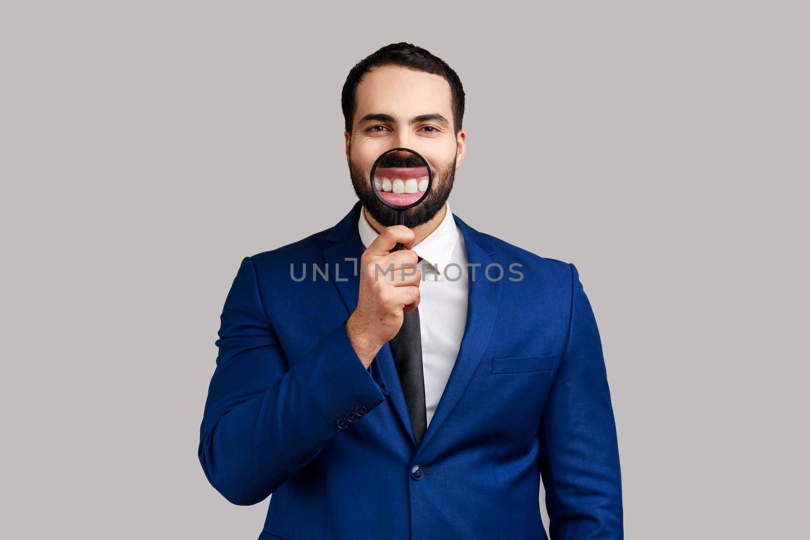 Portrait of bearded businessman holding magnifier on her teeth, looking at camera with happy expression, wearing official style suit. Indoor studio shot isolated on gray background.