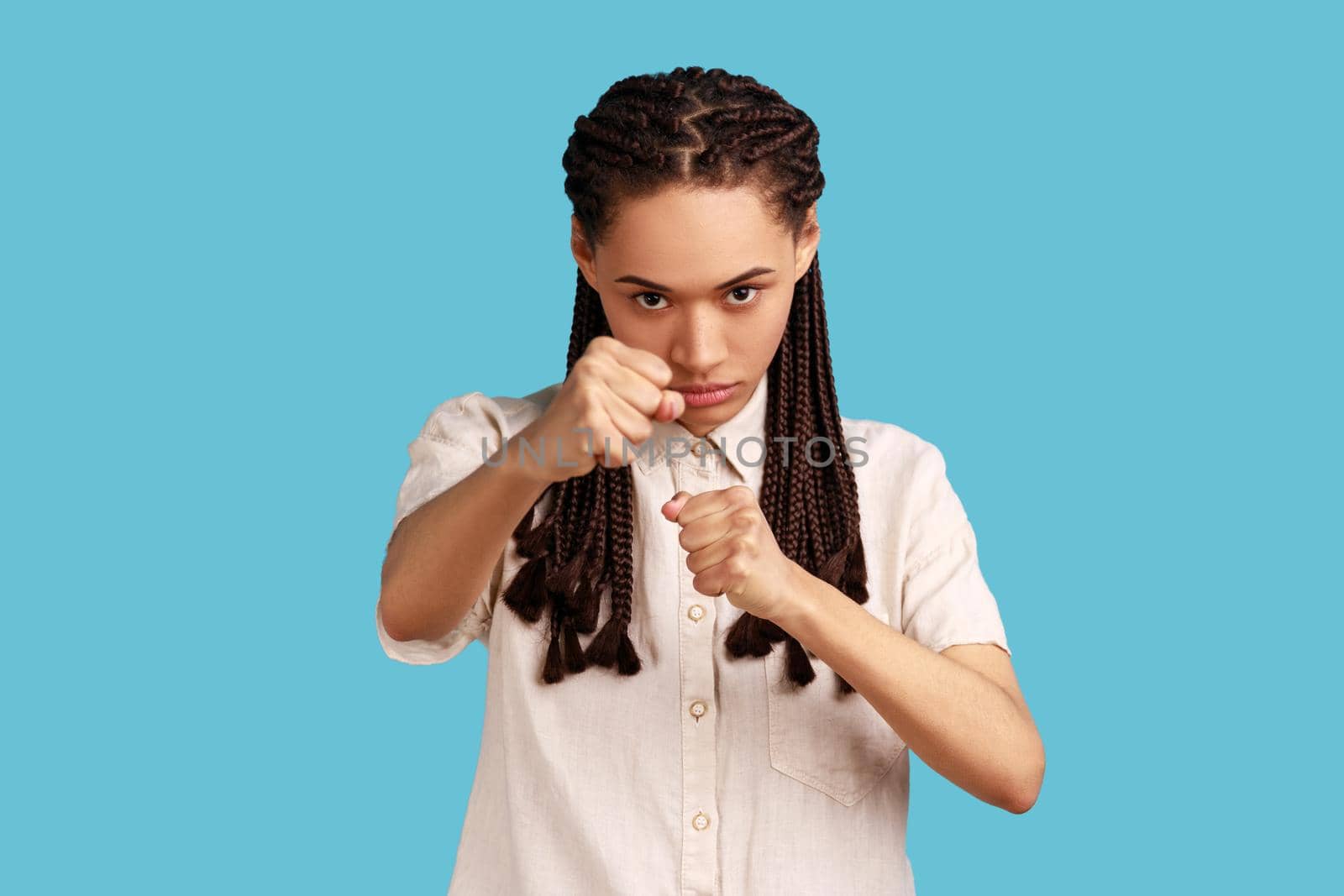 Woman with black dreadlocks having serious and confident look, keeping fists clenched in front of her, defencing herself from offence and bad treatment. Indoor studio shot isolated on blue background.