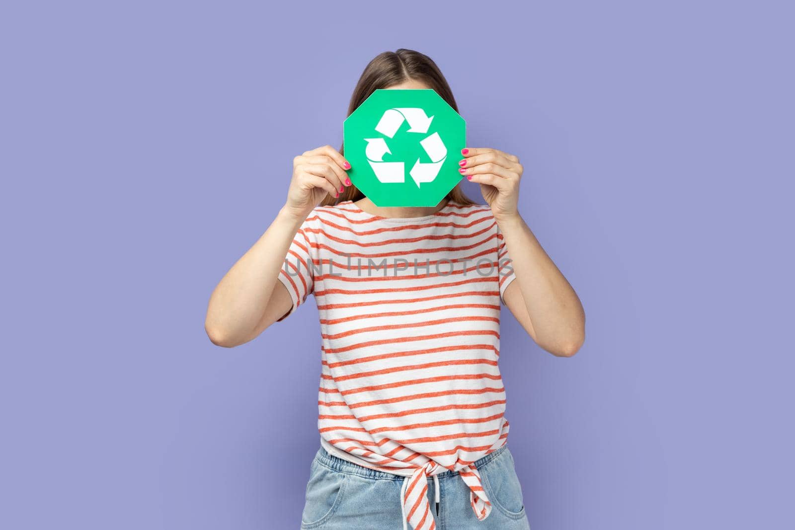 Portrait of unknown anonymous woman wearing striped T-shirt holding green recycling sign and hiding her face behind ecology symbol. Indoor studio shot isolated on purple background.