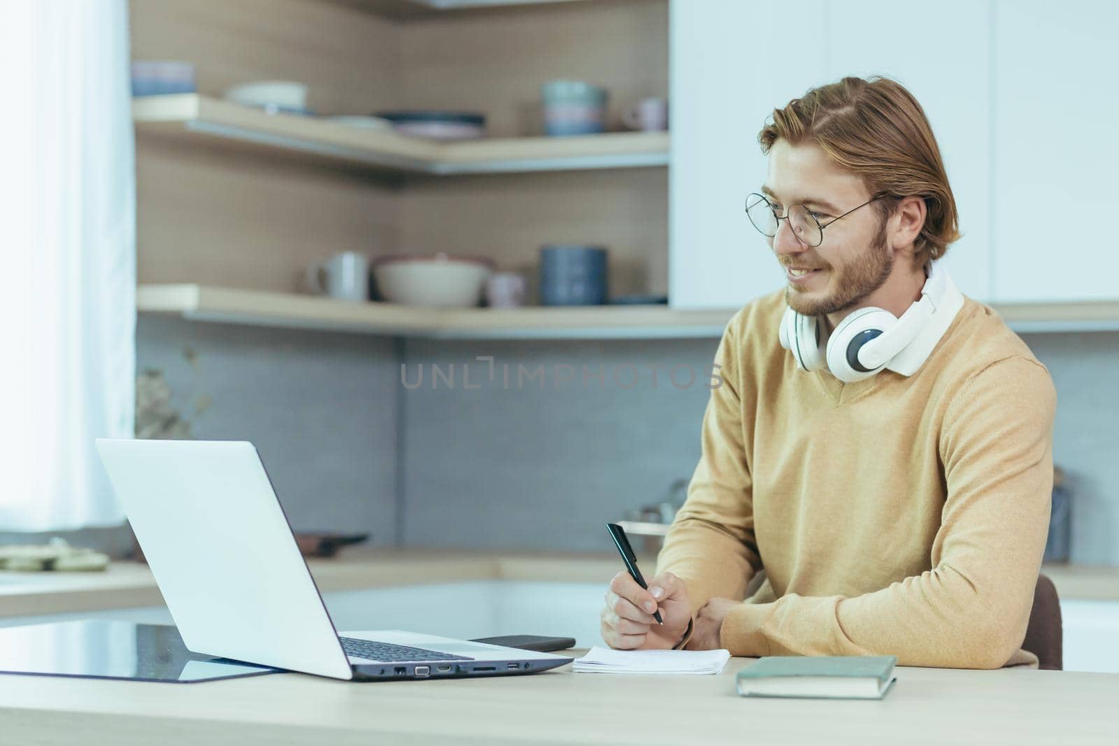 Young male entrepreneur working at home on paper work, sitting in the kitchen at the table, using a laptop for remote work as a freelancer.