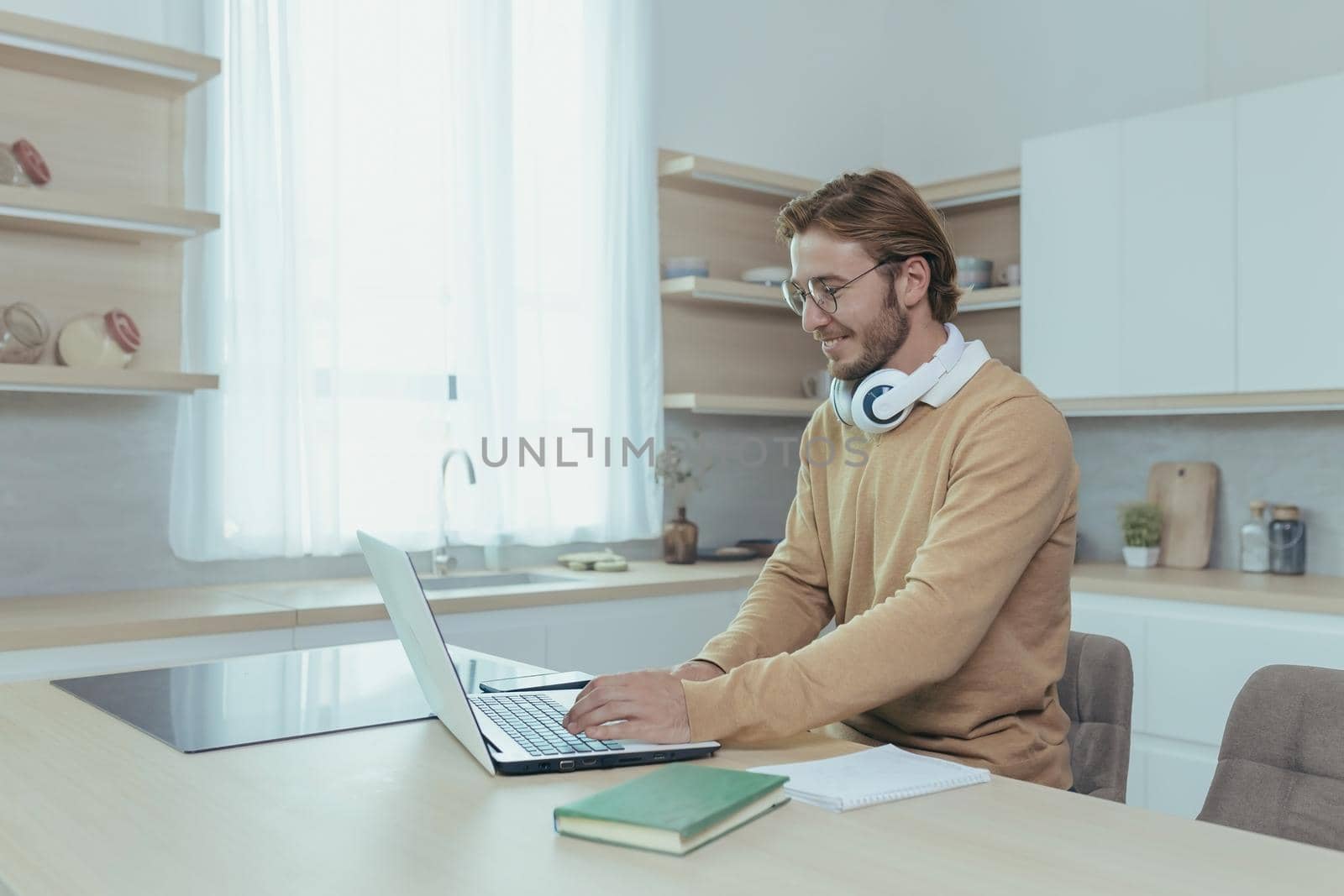 Young successful and happy businessman working from home, man using laptop for remote work, freelancer smiling in casual clothes sitting in kitchen at table.