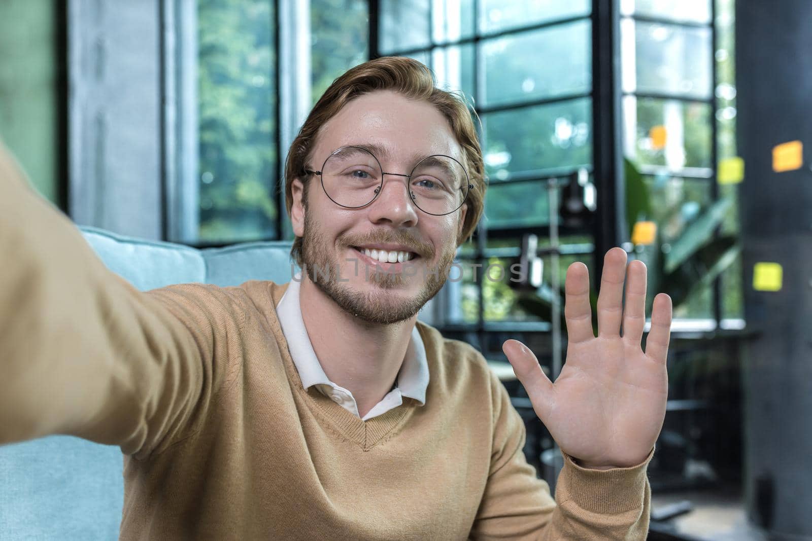 Man working in modern office, happy blond businessman smiling and looking at smartphone camera, holding hand up greeting gesture, worker in glasses taking selfie, video call with friends