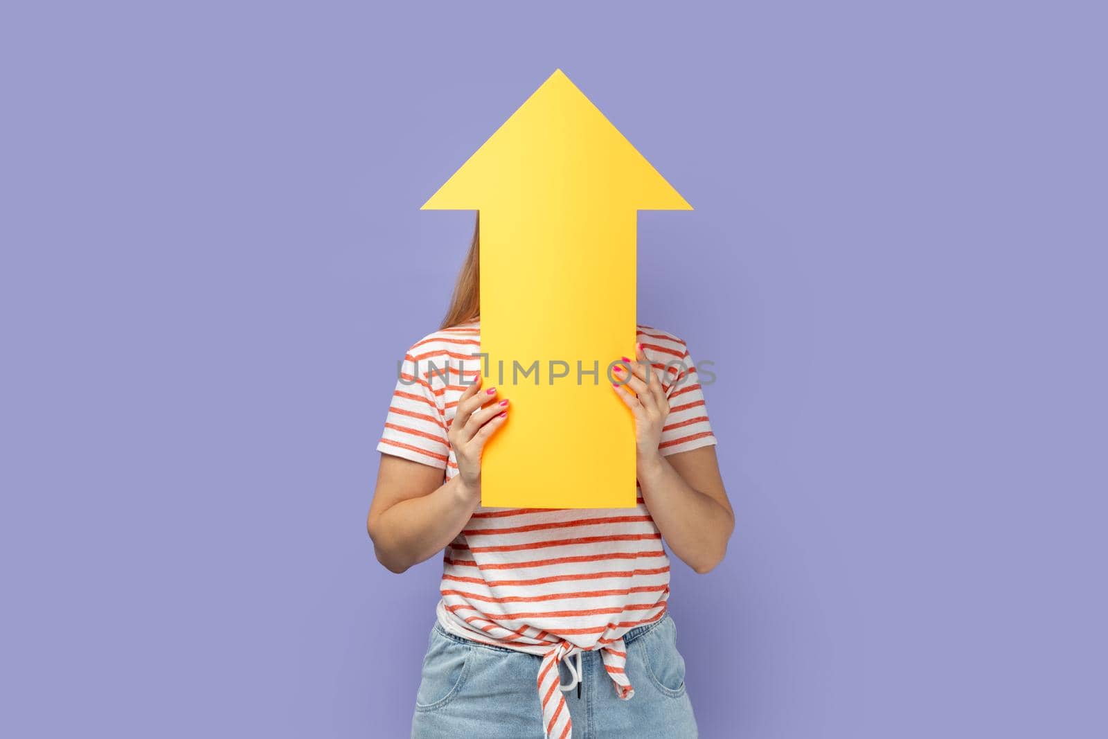Portrait of anonymous unknown blond woman wearing striped T-shirt holding big yellow arrow pointing up, showing increase in career. Indoor studio shot isolated on purple background.