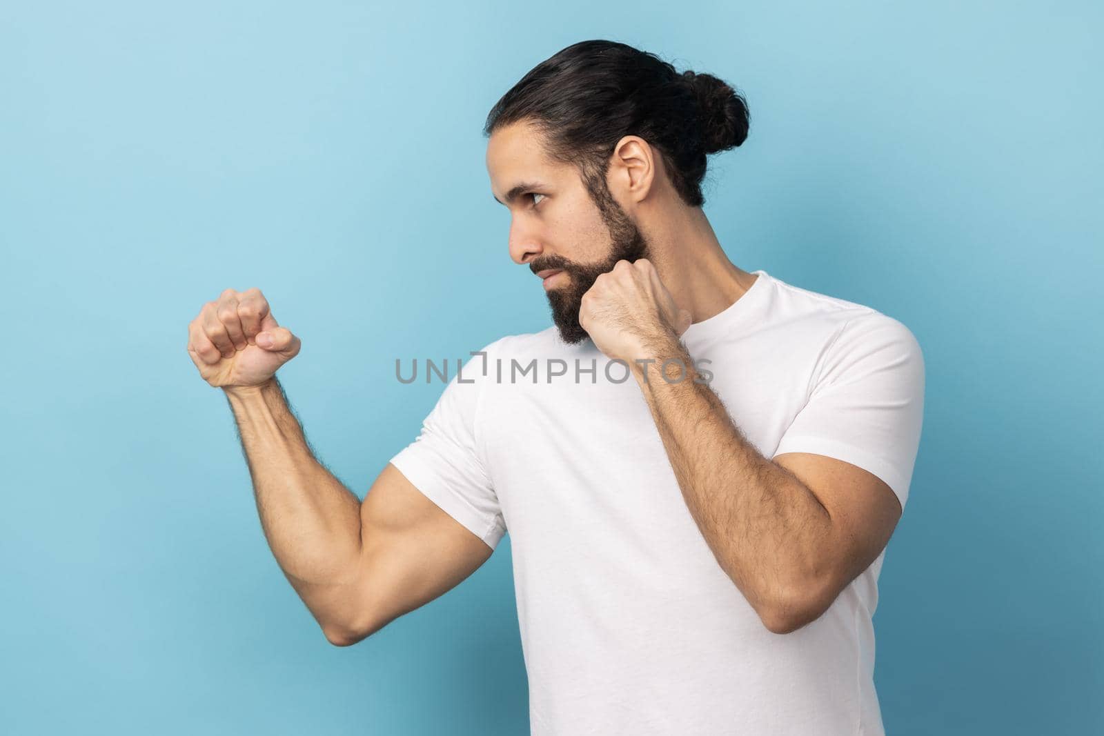 Side view of aggressive man with beard wearing white T-shirt holding clenched fists up ready to boxing, martial art trainer, self defense. Indoor studio shot isolated on blue background.