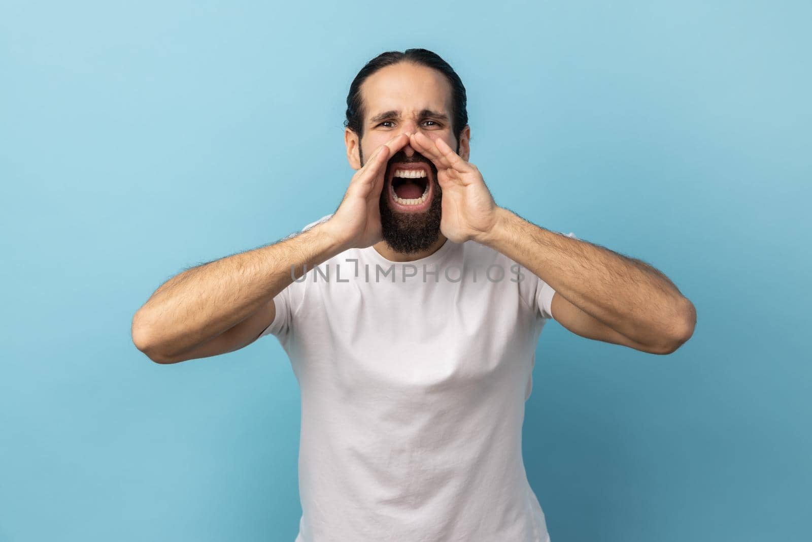 Attention, announcement. Portrait of handsome man with beard wearing white T-shirt screaming message, loud advertising, keeps hands near mouth. Indoor studio shot isolated on blue background.