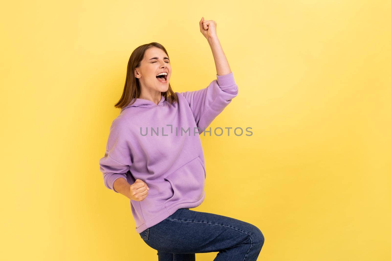 Portrait of enthusiastic woman screaming for joy and raising high her hand, celebrating successful winning, incredible victory, wearing purple hoodie. Indoor studio shot isolated on yellow background.