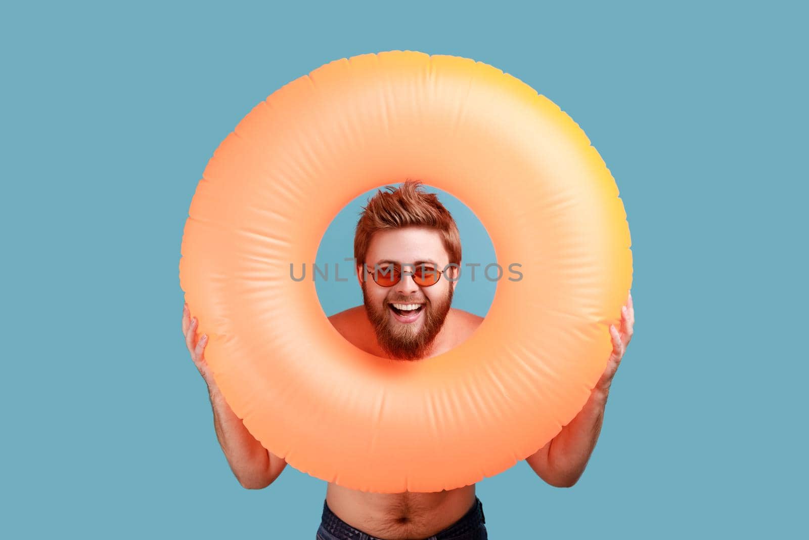 Portrait of bearded man in sunglasses holding orange rubber ring in hands, looking at camera with happy expression, enjoying tour. Indoor studio shot isolated on blue background.