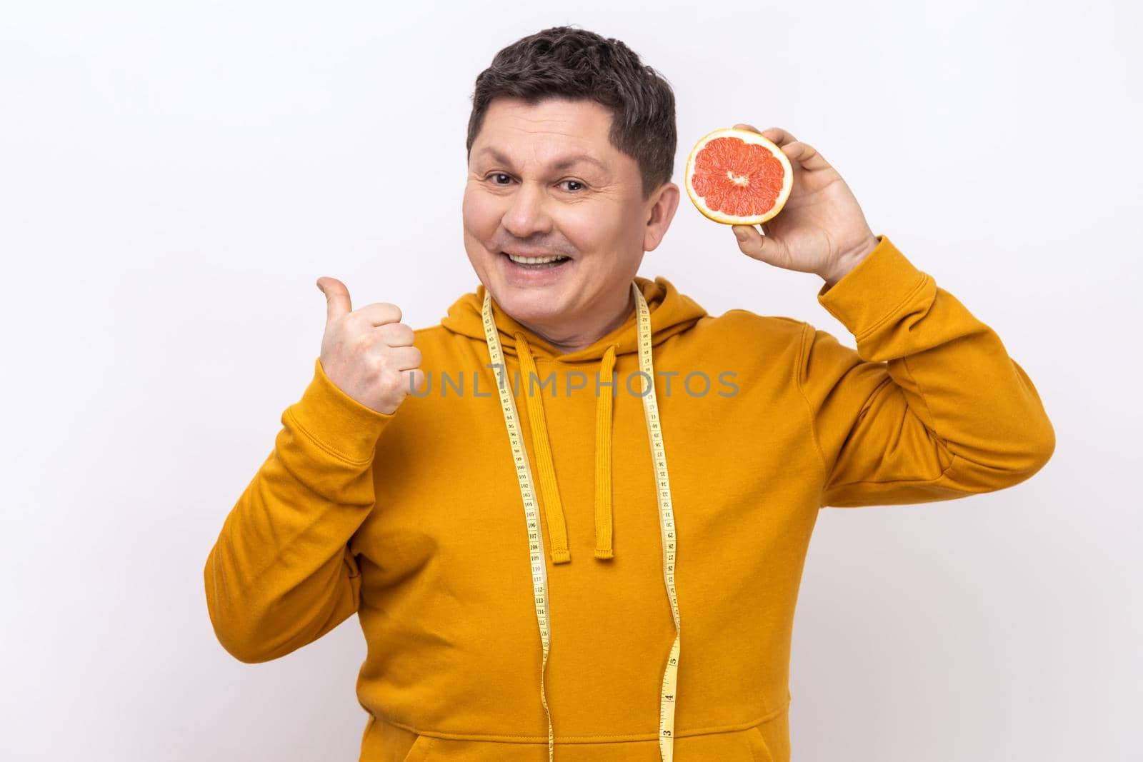 Middle aged man with dark hair holding slice of grapefruit, looking at camera with toothy smile, showing thumb up, wearing urban style hoodie. Indoor studio shot isolated on white background.