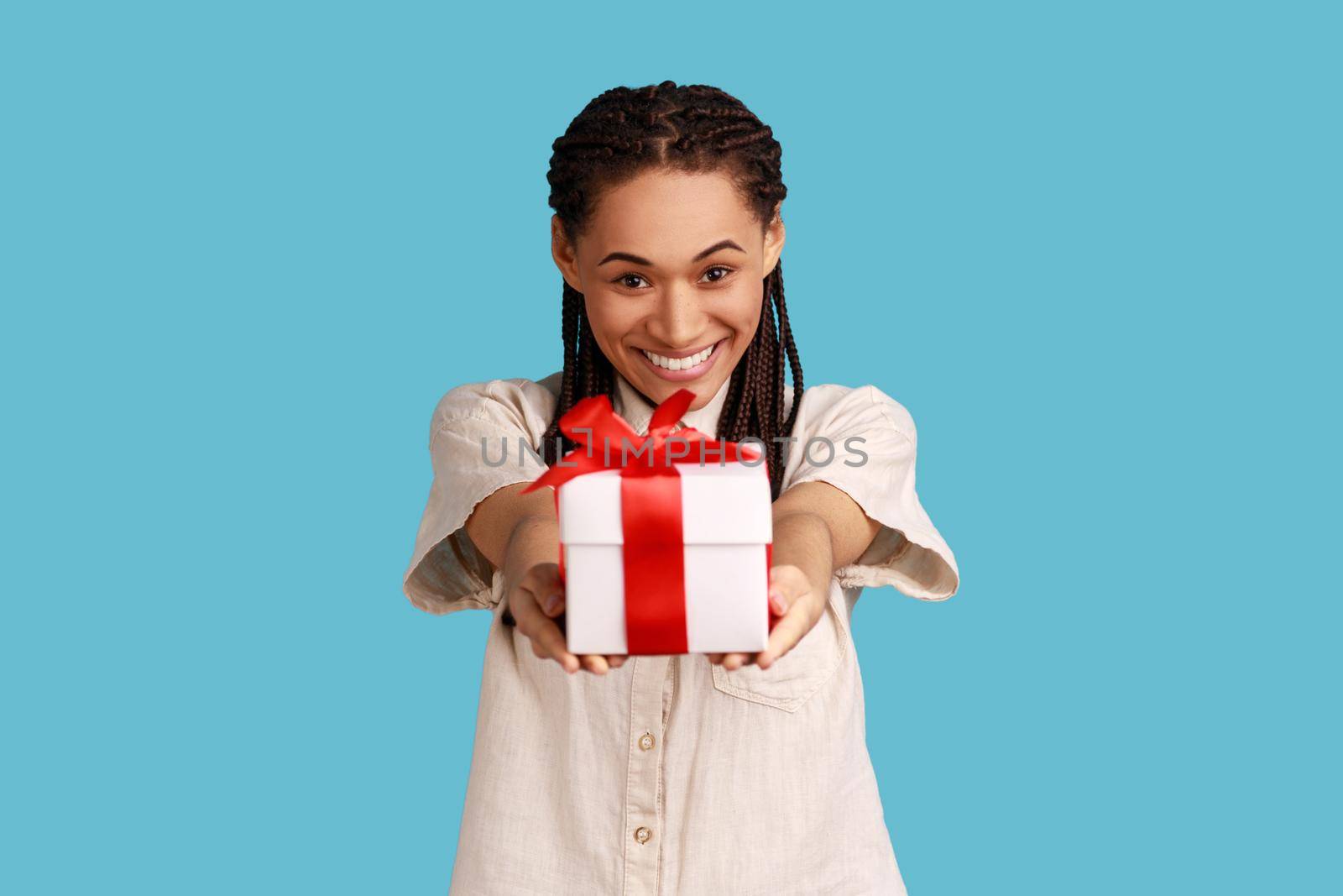Portrait of happy woman with black dreadlocks giving present box, looking at camera, congratulating friend with birthday, wearing white shirt. Indoor studio shot isolated on blue background.