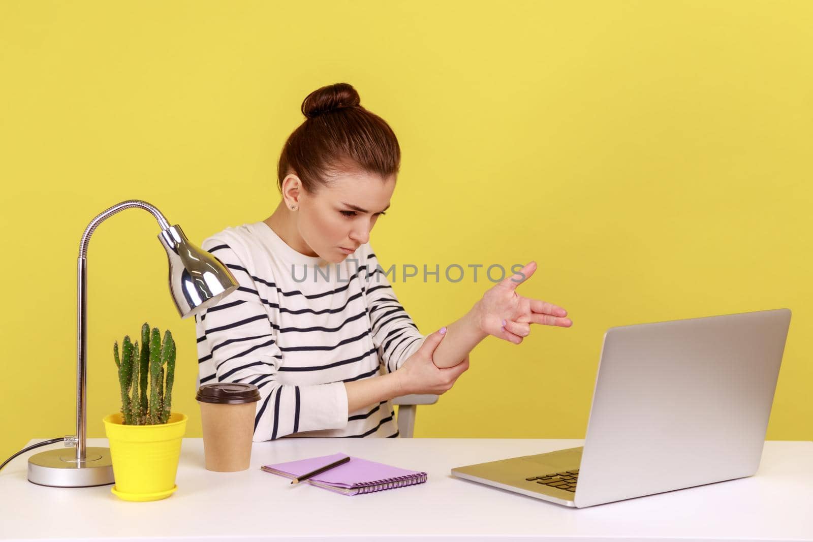 Aggressive woman manager sitting at workplace with laptop threatening and pointing finger gun to laptop screen, shooting with angry expression. Indoor studio studio shot isolated on yellow background.