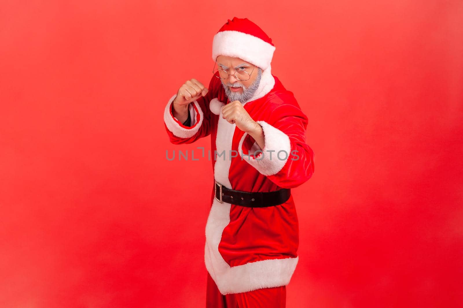 Strict elderly man with gray beard wearing santa claus costume standing with fists and being ready to fight, looking at camera with angry expression. Indoor studio shot isolated on red background.