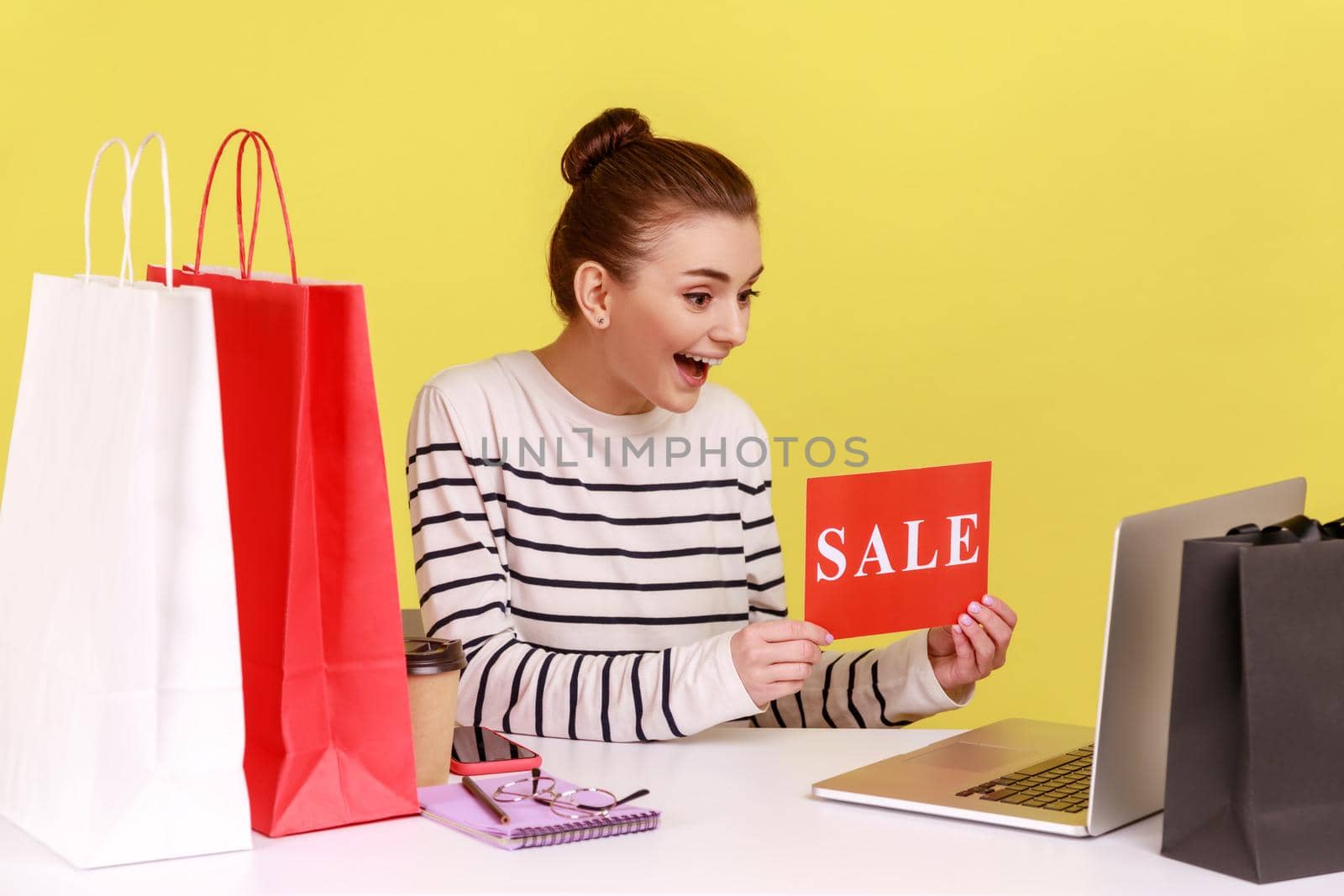 Excited woman office manager holding sale card looking at laptop monitor with happiness sitting at workplace with paper shopping bags. Indoor studio studio shot isolated on yellow background.