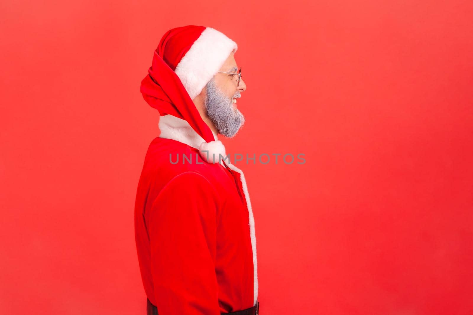 Side view of elderly man with gray beard wearing santa claus costume having positive expression, looking ahead, being in good festive mood. Indoor studio shot isolated on red background.