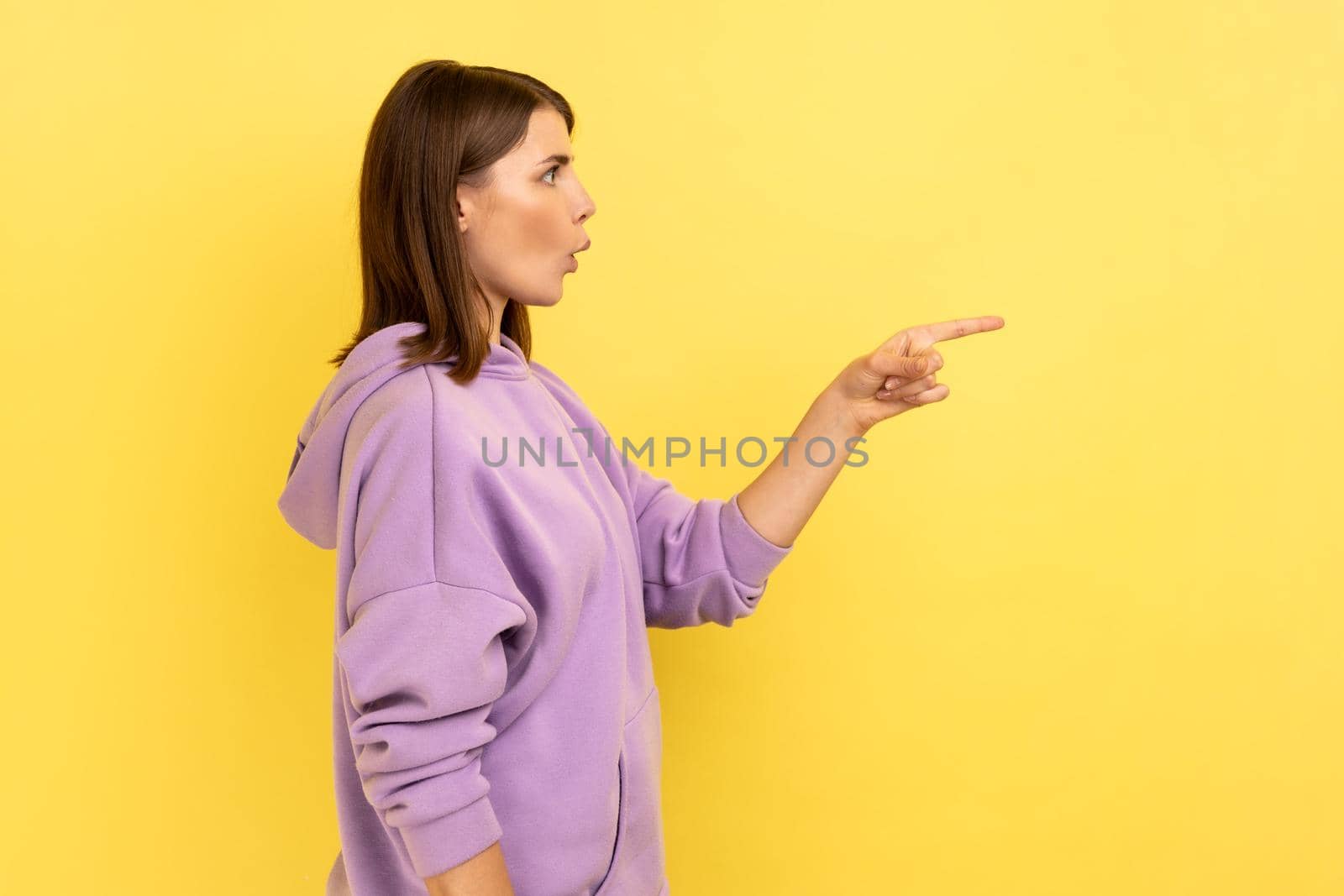 Side view of excited beautiful young adult woman standing and pointing at copyspace with surprised face, wearing purple hoodie. Indoor studio shot isolated on yellow background.