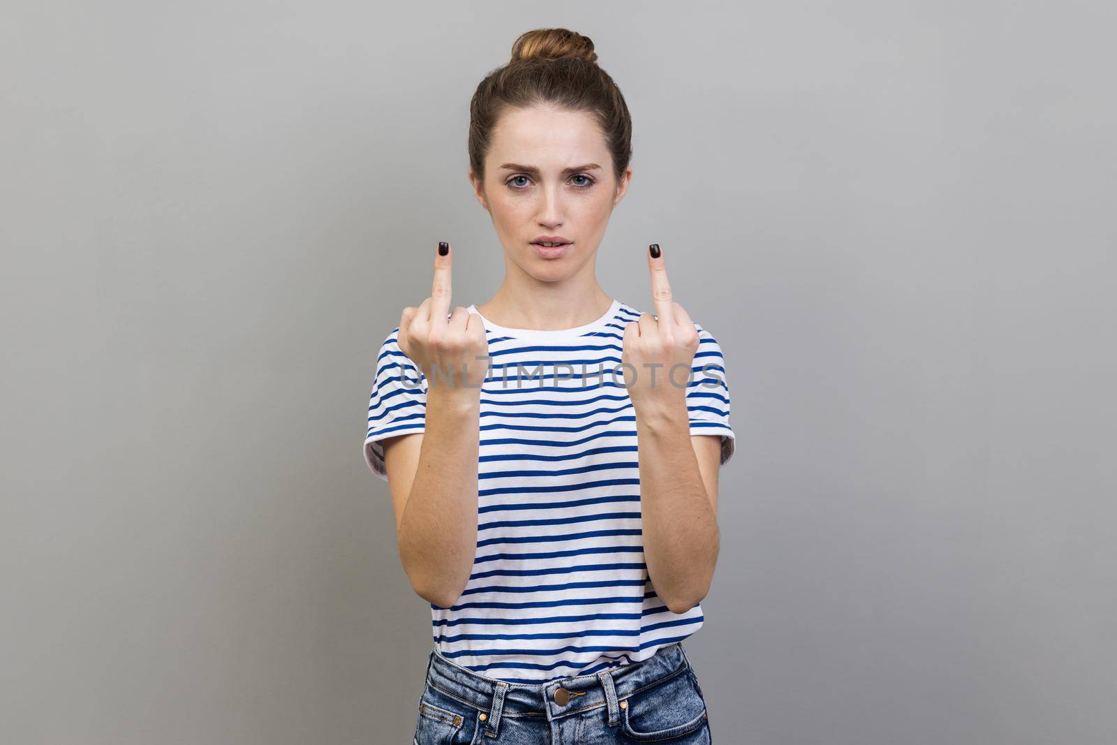 Portrait of crazy angry woman wearing striped T-shirt showing middle finger, rude gesture, face full of hatred and resentment. Indoor studio shot isolated on gray background.