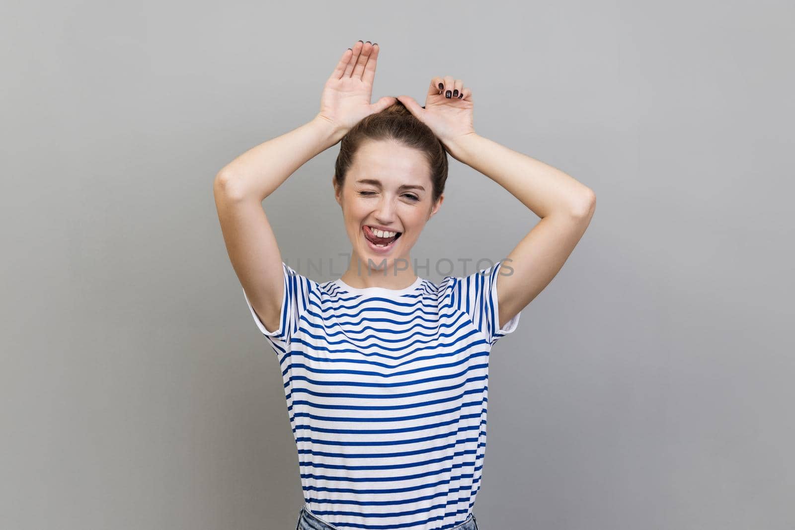 Portrait of playful woman wearing striped T-shirt making funny bunny ears with hands on head, childish behavior, optimistic mood. Indoor studio shot isolated on gray background.